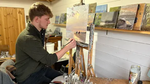 Qays Najm/BBC Kieron Williamson in his art studio, adding touches to a painting that shows two horses being ridden on a beach. He is wearing a green jumper and black trousers. On a shelf are various paintings he has produced.